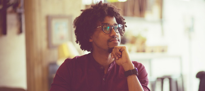 A young man with medium complexion and mid-length slightly locked hair and neat goatee wearing glasses and casual maroon shirt sits thoughtfully at a desk with a laptop, papers, and a smartphone in front of him, resting his chin on his hand and gazing into the distance in a warmly lit, casual workspace.