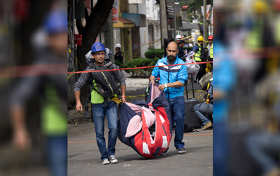 Two men, one wearing a blue hardhat and a stuffed backpack that he carries in front and the other bald with a dark beard and wearing a blue windbreaker with a black and white stripe across the front, carry a red, blue and pink blanket between them that is apparently filled with belongings as they walk through a damaged street in Mexico City, with men in hardhats working behind them, after the September 2017 earthquake