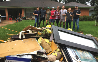 A group of eight students from the Jones School, all in work clothing, stand holding cans of soda on a lawn in front of a damaged suburban house and behind a large pile of debris that they have cleaned up from a neighborhood after Hurricane Harvey.