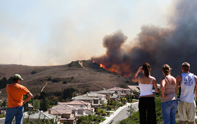Several people, including one woman in ponytail and orange shirt to the left and three people including two women and aman dressed in summer clothing, looking over a row of similar California suburban houses as a fire burns and smokes on a charred hill in the distance, with smoke obscuring the otherwise blue sky
