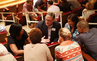 Marketing professor Ron Wilcox sits in a group of five people, including four women and a man all of different ethnicities, as they all listen intently to a Black woman speaking, with an open-hand gesture, during an August 14, 2017, community conversation.