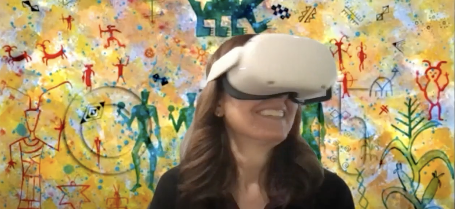 smiling woman wearing Oculus virtual reality glasses and standing before a colorful wall with Native American drawings on it.