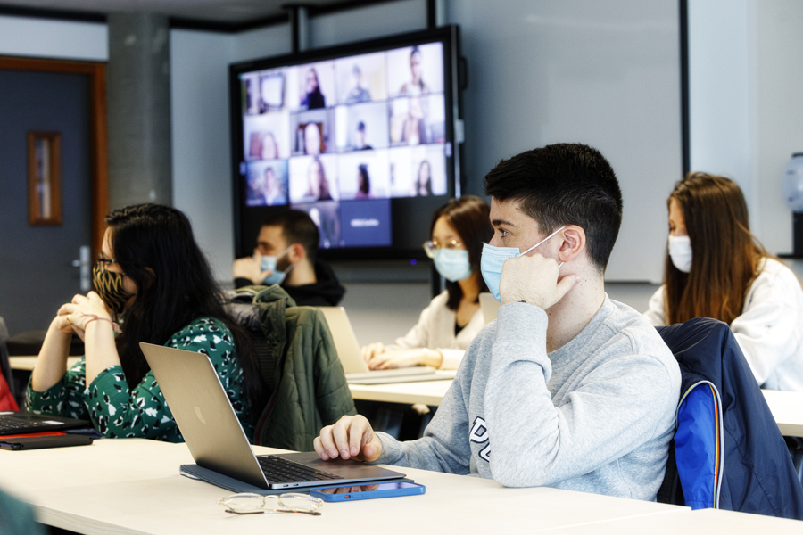 Students sit masked in a physical classroom with a large screen in the background displaying students logged in remotely in a Hyflex class at Grenoble Ecole de Management