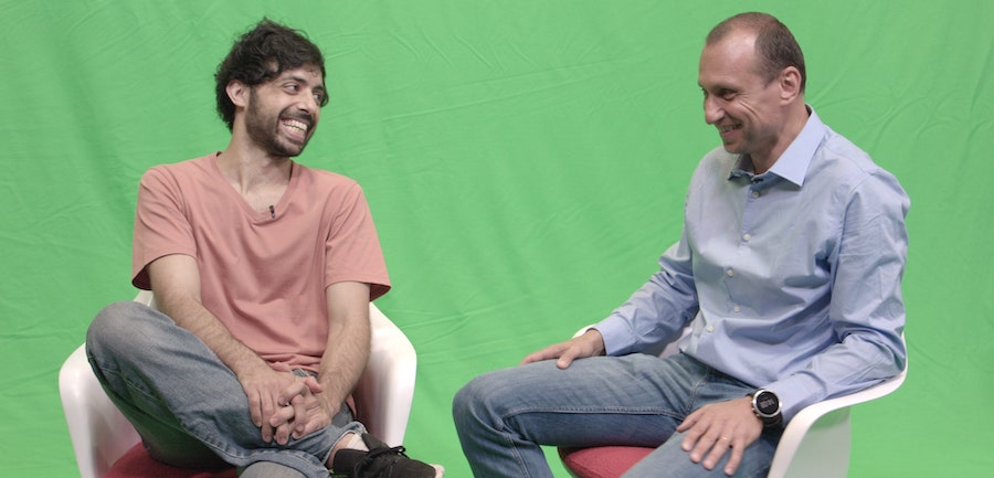 Two men sitting in chairs against a green screen, laughing