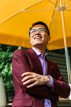 Dreen Yang of Coca-Cola wearing glasses with short dark hair, a burgundy blazer over a lilac button down with open collar, arms crossed, big smile, looking up to the left, all under a bright yellow patio umbrella