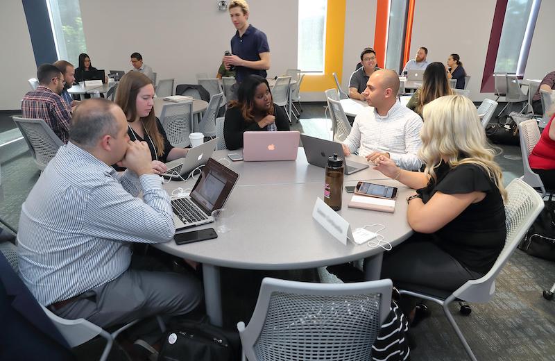 students sitting around a round table in a spacious room, looking at laptops and holding a discussion
