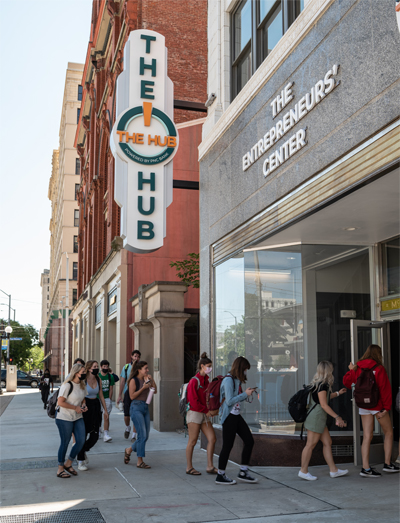 Students walk from the sidewalk into the large glass front doors of The Hub, a gray brick building with "The Entrepreneurs' Center" in white letters above the doors and a large vertical white marquee extends from the building that spells "The Hub" in vertical letters with a green circle inside which have the words "The Hub" in smaller orange letters