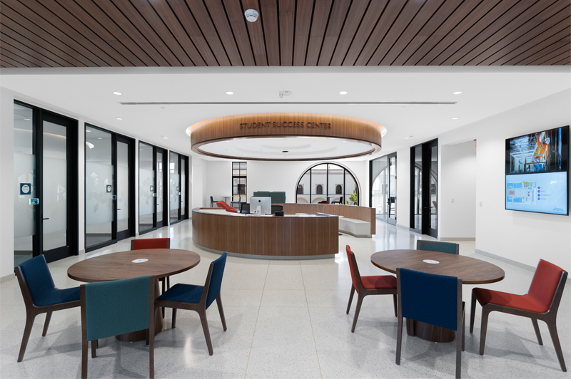 The Free Enterprise Institute at the Knauss School of Business at the University of San Diego, a large room with blue carpet, three short rows of computer stations in front of a projector screen, with two glass doors on either side facing the atrium outside, while the open area of the room contains three comfortable blue high-backed chairs and small wood and metal side tables, as well as a long white island of contemporary design with high stools, backed by a wall of windows that look into a breakout room