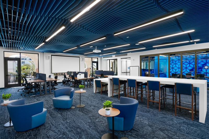 The Free Enterprise Institute at the Knauss School of Business at the University of San Diego, a large room with blue carpet, three short rows of computer stations in front of a projector screen, with two glass doors on either side facing the atrium outside, while the open area of the room contains three comfortable blue high-backed chairs and small wood and metal side tables, as well as a long white island of contemporary design with high stools, backed by a wall of windows that look into a breakout room