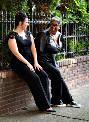 Tippie College's Beth Livingston with light skin, dark hair upswept into a loose bun and wearing a black shirt with lace cap sleeves with dark pants sits with Babson's Tina Opie with dark complexion and glasses, short salt-and-pepper hair, wearing a chunky gold necklace and a leather shirt with deep front-V and loose three-quarter sleeves and dark pants laugh with one another as they sit on the narrow ledge of a half-brick wall leaning back against wrought iron fencing with greenery coming through around them
