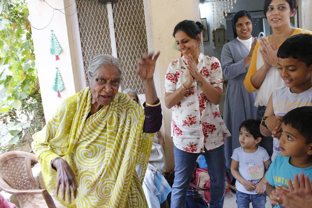 Simran Oberoi stands smiling and clapping as an elderly woman in a beautiful light yellow sari dances to the left of her and two workers and three children stand to the right and behind watching and smiling
