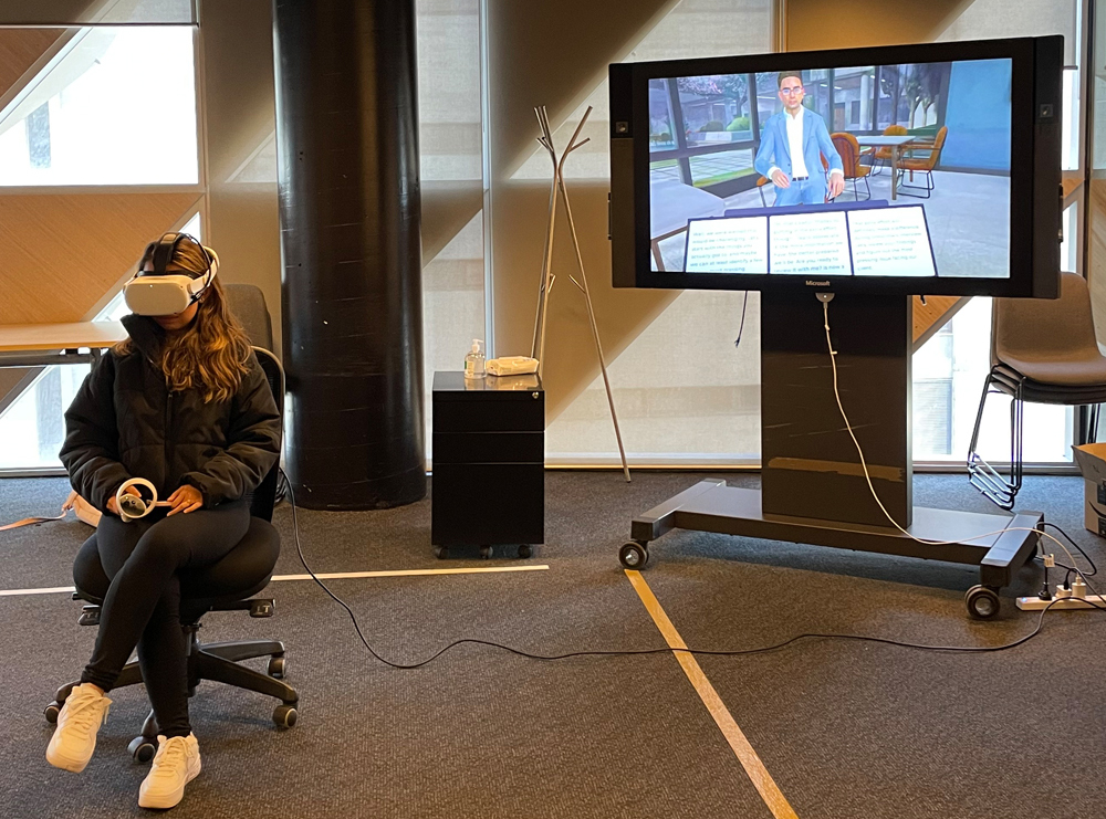 In a large spacious room with gray carpeting and high ceilings, a young woman with long wavy brown hair and wearing a long-sleeve zip-up jacket, leggings, and white sneakers wears a VR headset and holds VR hand controllers as she sits in an office chair, legs crossed, to the left with a flat-screen display to the right and slightly behind her showing the VR-simulated male client she is engaging with.