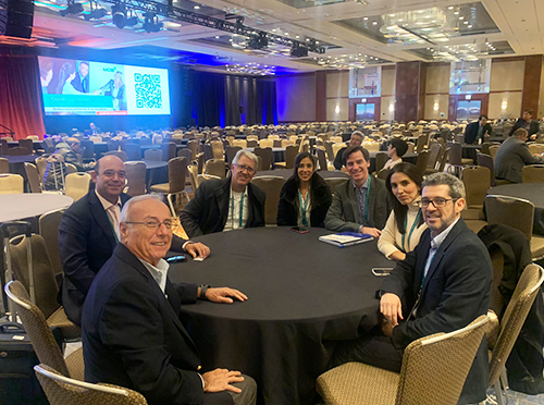 Photo of seven people sitting at a round table in a large conference room that is otherwise empty, smiling at camera. ICAM 2023