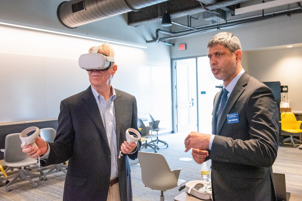 A college professor stands in a classroom with an older man who is wearing virtual reality goggles; the professor is explaining how virtual reality works.