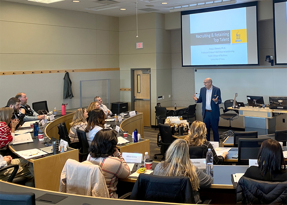 An executive education classroom, seen from the back, with students of all types seated in curved rows and the professor gesturing and talking in the front of the class in front and to the left of a light-wood-colored table-like lecturne with his laptop and monitor on it. Behind the professor and lecturne is an empty whiteboard and above that is a large projection screen on which a PowerPoint slide reads "Recruiting and Retaining Top Talent" 