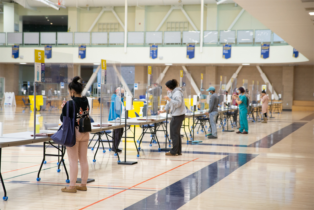 Students and staff at UC Davis stand at tables, placed six feet apart, to register for COVID testing at a testing center set up in the school's basketball arena in 2020