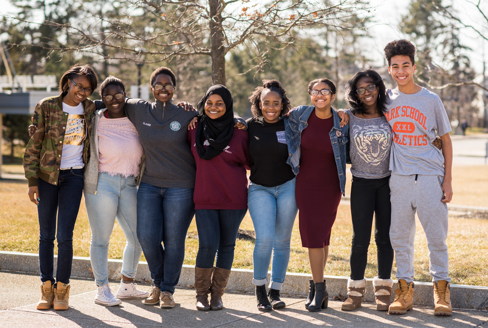 Eight Buffalo Prep students stand in a line together, smiling widely and facing the camera with their arms around each other's shoulders and waists, on the south campus of the University at Buffalo. The six young women and one young man(at far right) stand on a sidewalk with the sun behind them casting their shadows before them, with a yellowing lawn, trees with a few leaves of autumn left on their branches, and a gray-blue sky in the background. From left: Te’Aira Alexander ’15, Maria Sibomana ’13, Shamso Osman ’13, Khaliah Beaver ’13, Taylor Lewis ’15, Mary Wright ’15 and Angel Leon ’16. Photo: Dawn M. Gibson