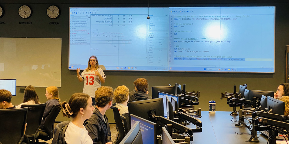 Professor Kathryn Bender, wearing a white short-sleeved sports-style jersey with "Data Enchanted" and "13" printed on it in red, teaches in front of a classroom of students sitting at long tables, positioned perpendicular to the front of the room, with monitors on swing arms in front of them. Behind and above Bender is a very large screen displaying programming code.