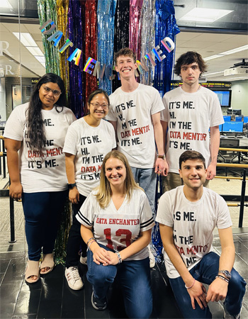 Professor Kathryn Bender with five Data Mentors that support her "Data Enchanted" data analysis workshop series. Bender crouches one knee down on the floor next to a young man doing the same, as four other mentors--two young women and two young men--stand behind them. The student data mentors all wear white t-shirts with the words "It's Me. I'm the Data Mentor. It's Me." a reference to Taylor Swift's song, Anti-Hero. Bender wears a white sports-style jersey t-shirt with the words "Data Enchanged" over the number 13, all in red. The entre group is gathered before the windows of the classroom with colorful metallic vertical streamers behind them and a string with colorful metallic letters spelling out "Data Enchanted" draped over the streamers.