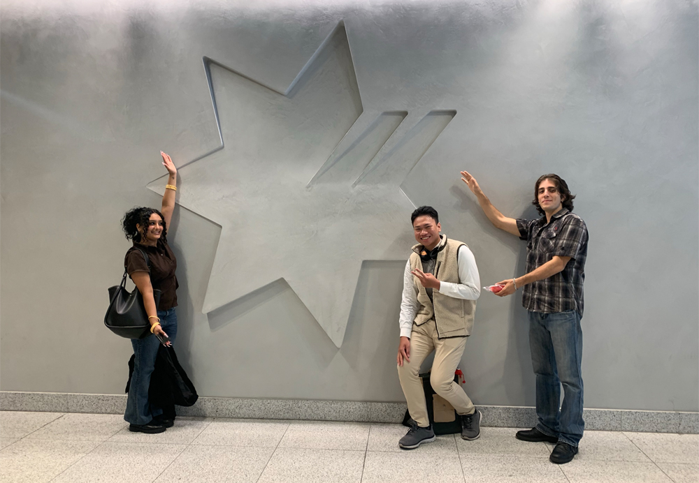 Three RMIT students stand smiling before a tall gray metal wall into which the seven-pointed star logo of the National Australia Bank is molded, with the top points carved into the metal while the bottom four points extend out in 3-D fashion. The young woman with long curly dark hair, black shirt and purse, and blue jeans stands at left with her left hair held up in the air to highlight the logo, while two young men stand to the right of the logo, one in khaki pants and a white long-sleeved shirt under beige vest stands crouched and showing a "peace" sign with his left hand and the other with long hair and wearing a black-and-gray plaid short-sleeve shirt and jeans stands with both arms extended as if gesturing toward the logo.