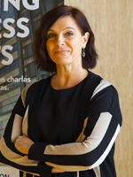 Katharine D'Amico of UPF Barcelona School of Management. She has a light complexion and slight friendly closed smile tilted slightly upward on one side, with dark eyes and brows and dark side-parted hair styled in a bob. She wears a black crew neck shirt with a white-stripe embellishment on each shoulder and drop earrings, and she stands in front of a light-wood paneled wall and an only partially visible promotional banner for the business school.