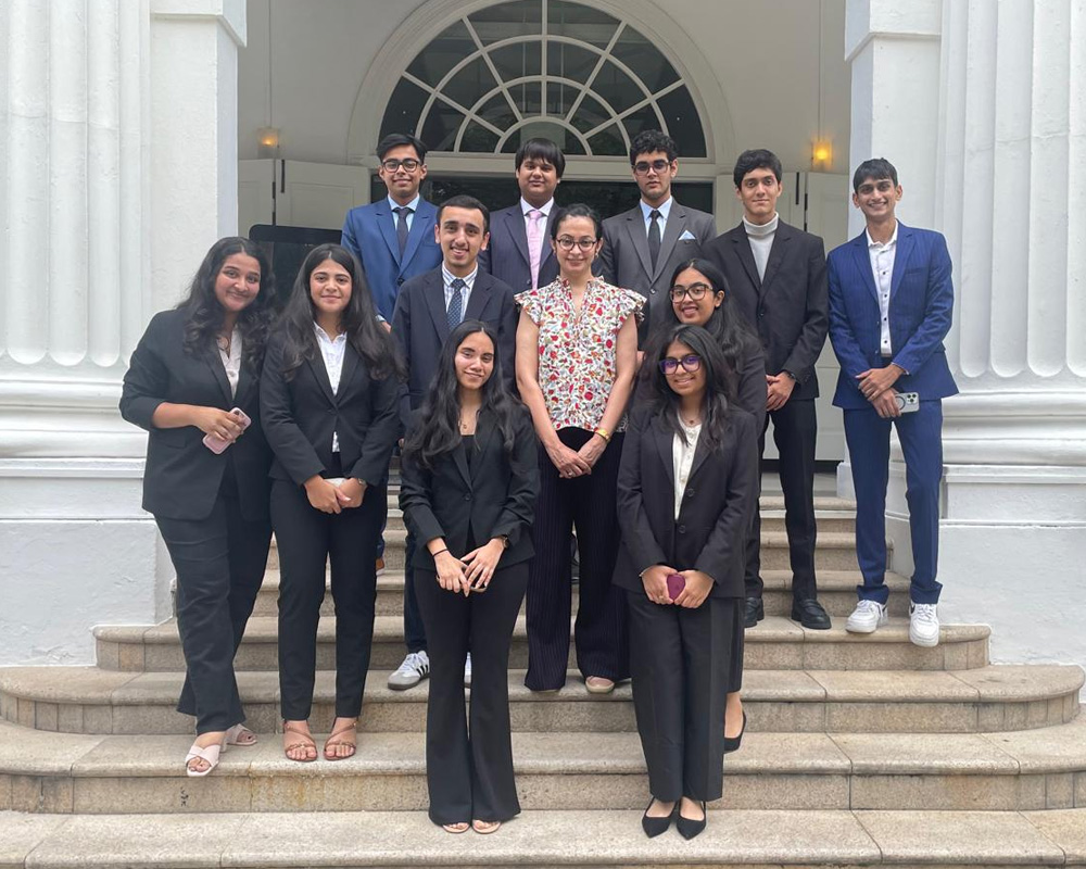 Professor Muniza Anzari of SP Jain School of Global Management stands center, dressed in a light floral short sleeved top and black pants, with 11 of her students standing around her, all dressed in business attire, on the cream-colored stone steps of a campus building. Large white columns flank them on either side, and the building's tall arched atrium window is visible behind them.