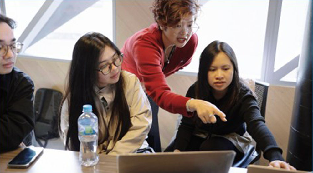 Instructor Tram Nguyen in a red cardigan stands behind three young adults who are looking intently as she leans over to point at the screen of the laptop on the table before them, with large windows in the background.