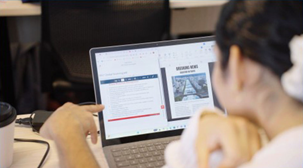 An over-the-shoulder view of a two students, both young women who are partially visible. One points at and one looks at a laptop screen displaying two open windows: a digital document on the left and a newsletter-style layout titled "Breaking News" on the right.