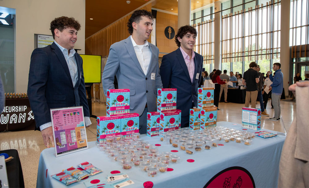 Three young men in suits, including Justin Ianelli, far left, and Harrison Nastasi, far right, stand at the Rowan Expo behind a light-blue covered table displaying boxes of their product, Bobica granola bars, as well as small sample containers of food, promotional signs, and nutrition information sheets. Other attendees and a bright atrium setting are visible in the background.