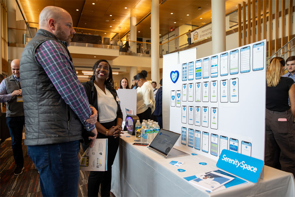 A young woman in a black jacket speaks to a man in a puffy vest and plaid shirt in front of a table with a display board describing her business idea, SerenitySpace. The board features a grid of smartphone mockups showing app screen designs, and on the table are water bottles, and brochures.