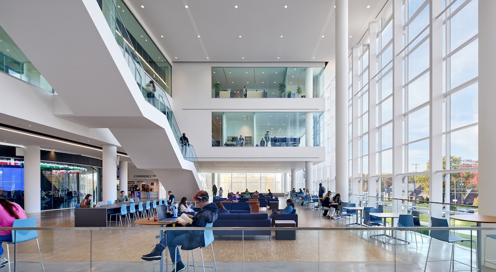 Huge open-air atrium with glass walls that extend several stories up on the right; on the left is a white staircase going up. Chairs and seating areas fill the floor but the impression is of light and air.