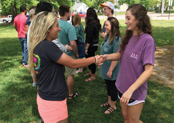 Photo of two young women outdoors wearing casual clothing, smiling and shaking hands, other students behind them. Courtesy of Tennessee Tech Marketing and Communications Department.