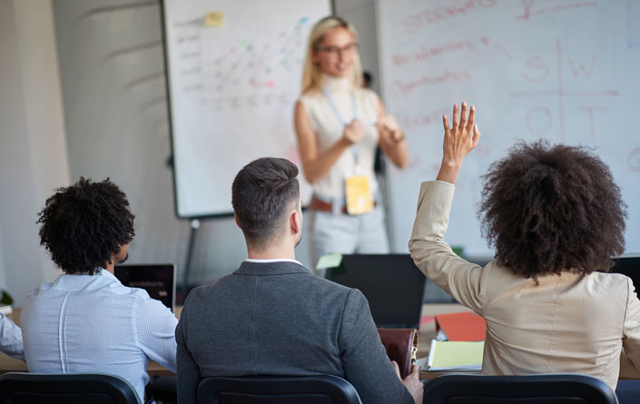 Three young professionals, a Black man, white man, and Black woman, are shown from behind as they sit in a classroom listening intently to the instructor, shown in soft focus in the background. The instructor is a slim young woman with straight shoulder-length blond hair, with glasses, a white sleeveless turtleneck, gray belted pants, and a lanyard around her neck with a yellow card hanging from it. The young Black woman, who is seated to the right and has shoulder-length natural curly hair and is wearing a beige suit jacket, has her left hand raised. The instructor is looking at her as if prepared to call on her and answer her question.