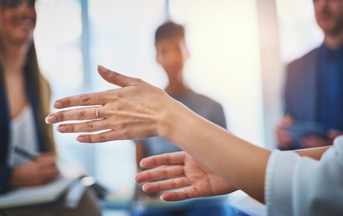 A close-up shot focuses on a woman's hands, a delicate gold ring on her middle finger, being held apart as if showing a gap. A bright office setting is visible in the blurred background, where three colleagues listen attentively, including a woman on the left taking notes.